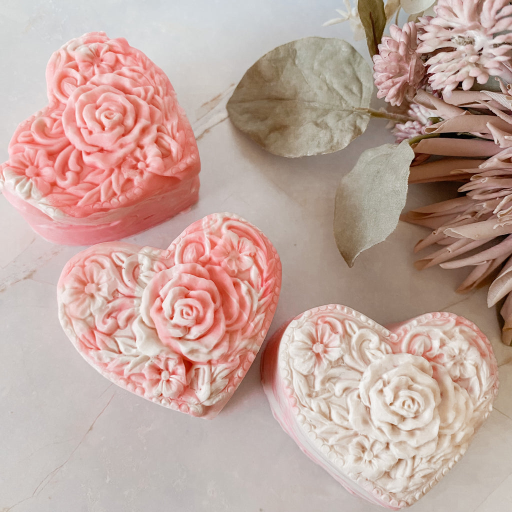 Three heart-shaped pink and white soaps with floral designs on a light surface with flowers and leaves.