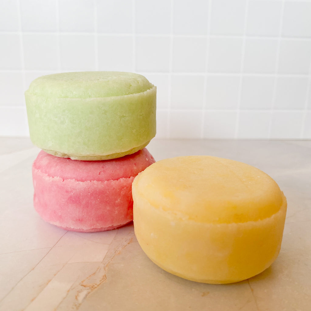Three colorfulshampoo bars stacked on a marble surface with a tiled wall background.