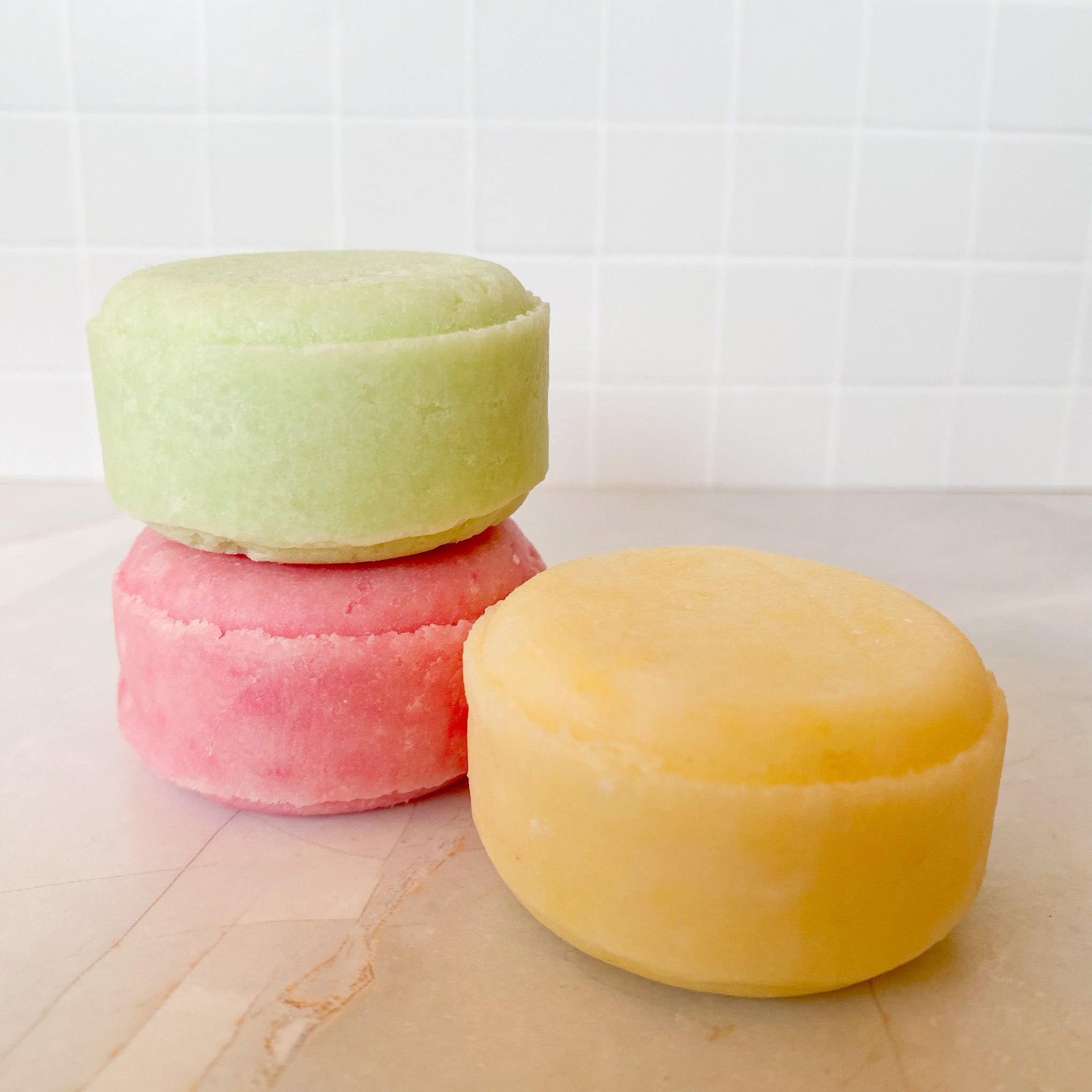Three colorfulshampoo bars stacked on a marble surface with a tiled wall background.