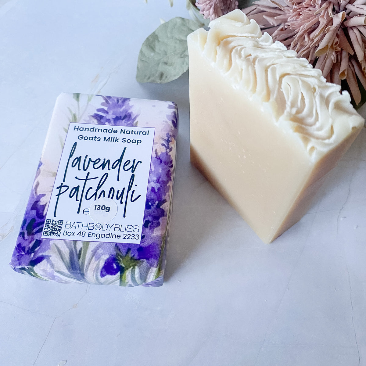 A bar of lavender patchouli handmade natural goat's milk soap shown as both wrapped packaging and an unwrapped cream-colored bar with swirled tops, styled with dried flowers on a light marble surface.