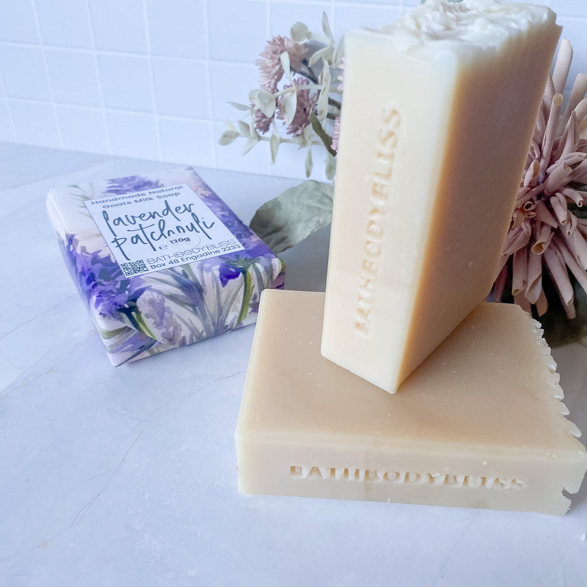 Two bars of Bath Body Bliss lavender patchouli soap on a light marble surface, with one bar standing upright and the other lying flat, alongside the branded wrapped soap package and soft floral decor.