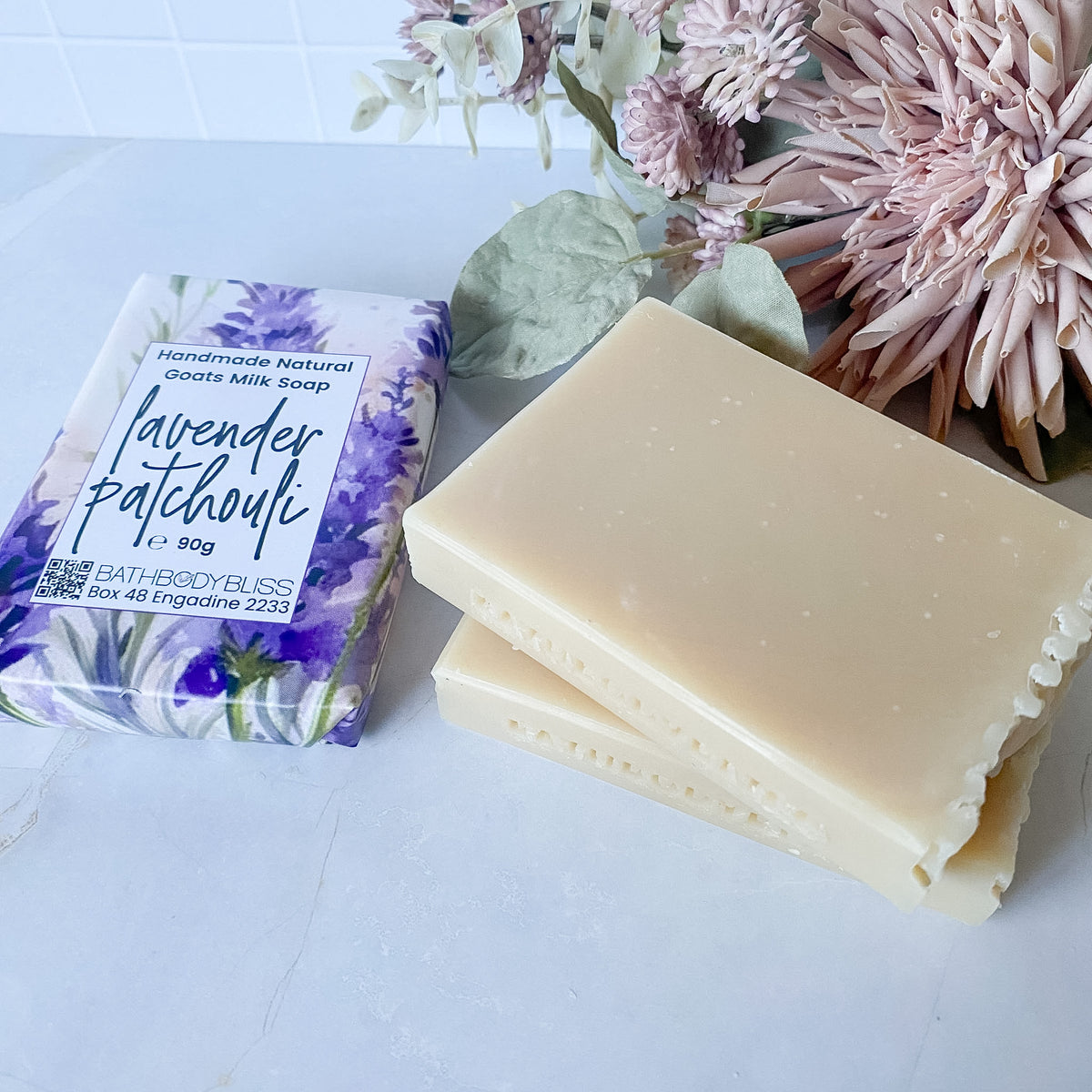 Three bars of Bath Body Bliss lavender patchouli goats milk soap, one wrapped in a purple floral paper sleeve and two unwrapped showing the creamy beige bar, styled with dried flowers on a white surface.