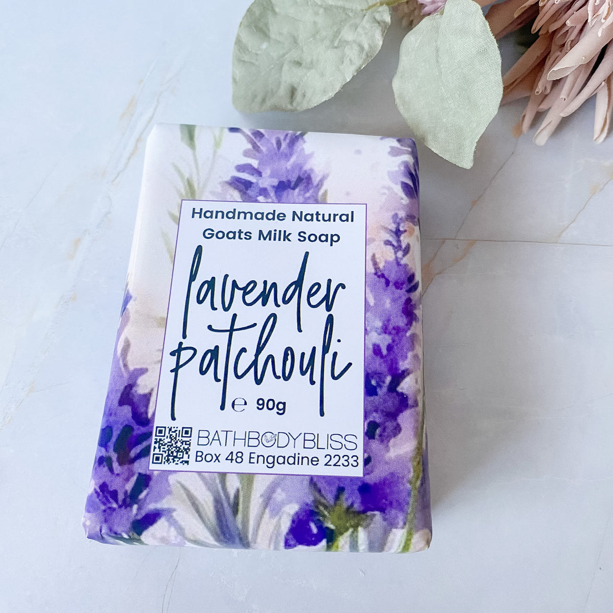 A 90g bar of lavender patchouli handmade natural goats milk soap by BathBodyBliss, wrapped in a watercolor purple and green floral paper sleeve, displayed on a light marble surface with dried leaves.