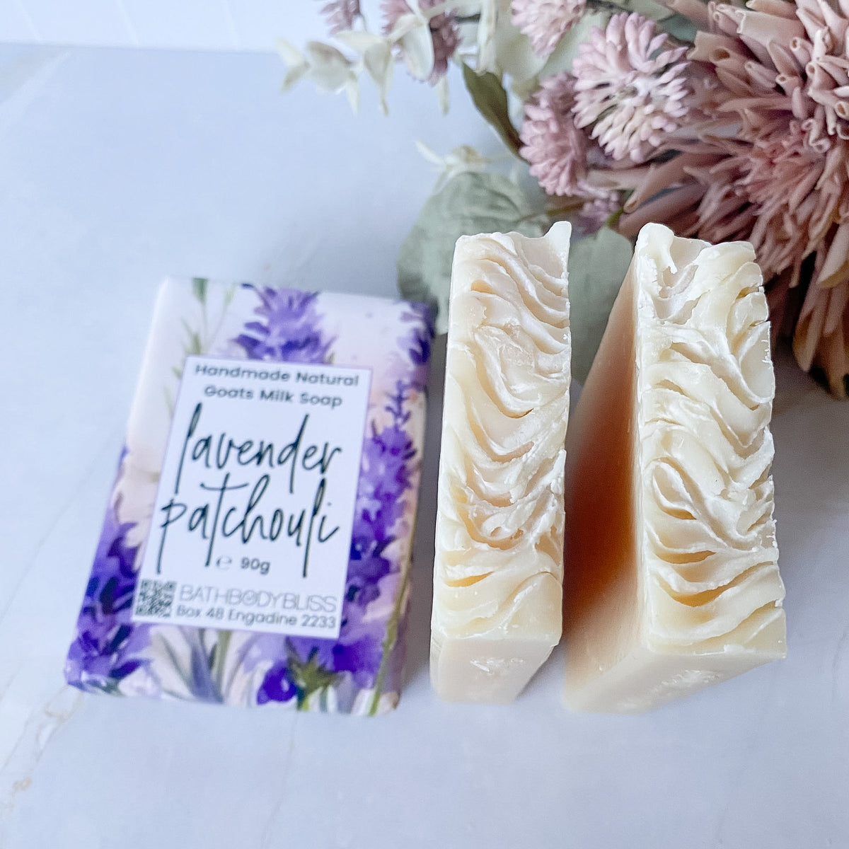 A bar of handmade soap shown as both wrapped packaging and unwrapped, alongside dried flowers on a light surface. The soap is labeled Bath Body Bliss Lavender Patchouli.