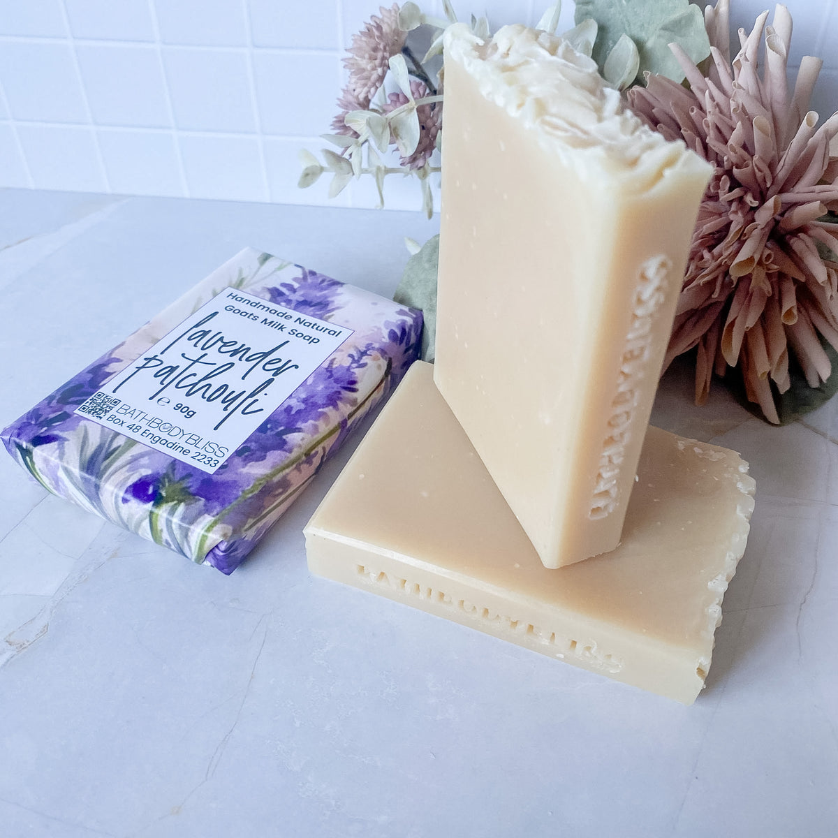 A bar of lavender-infused soap stands upright on another bar on a light marble surface, with the wrapped soap package beside it and dried flowers in the background.