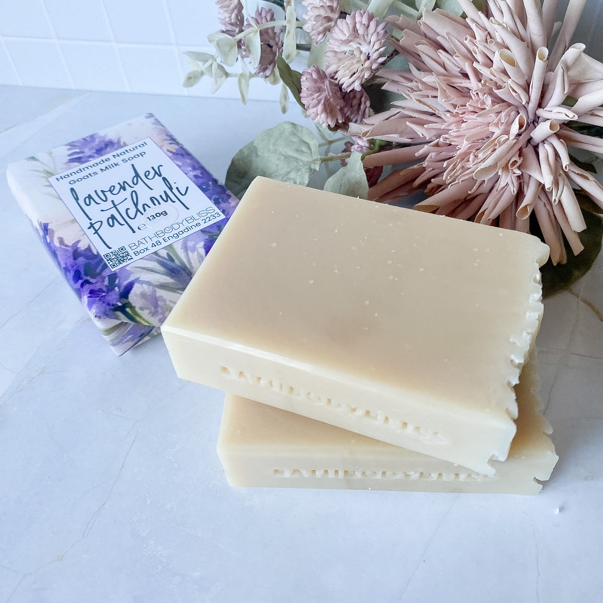 Two rectangular bars of pale cream soap stacked on a light surface, with a matching lavender patchouli scented soap bar wrapped in paper packaging behind them and dried pink flowers in the background.