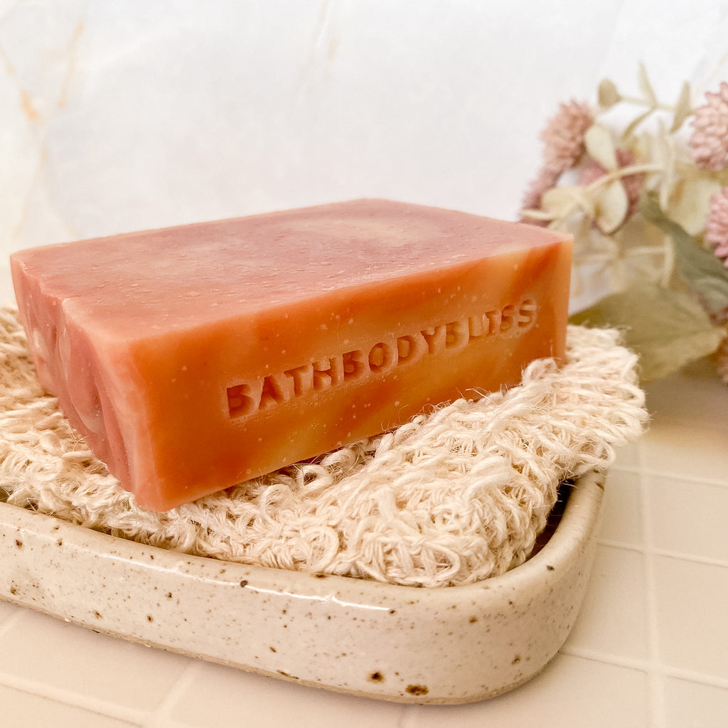 Bar of pink swirled soap with 'BathBodyBliss' branding on a natural fibre soap saver.