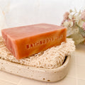 Bar of pink swirled soap with 'BathBodyBliss' branding on a natural fibre soap saver.