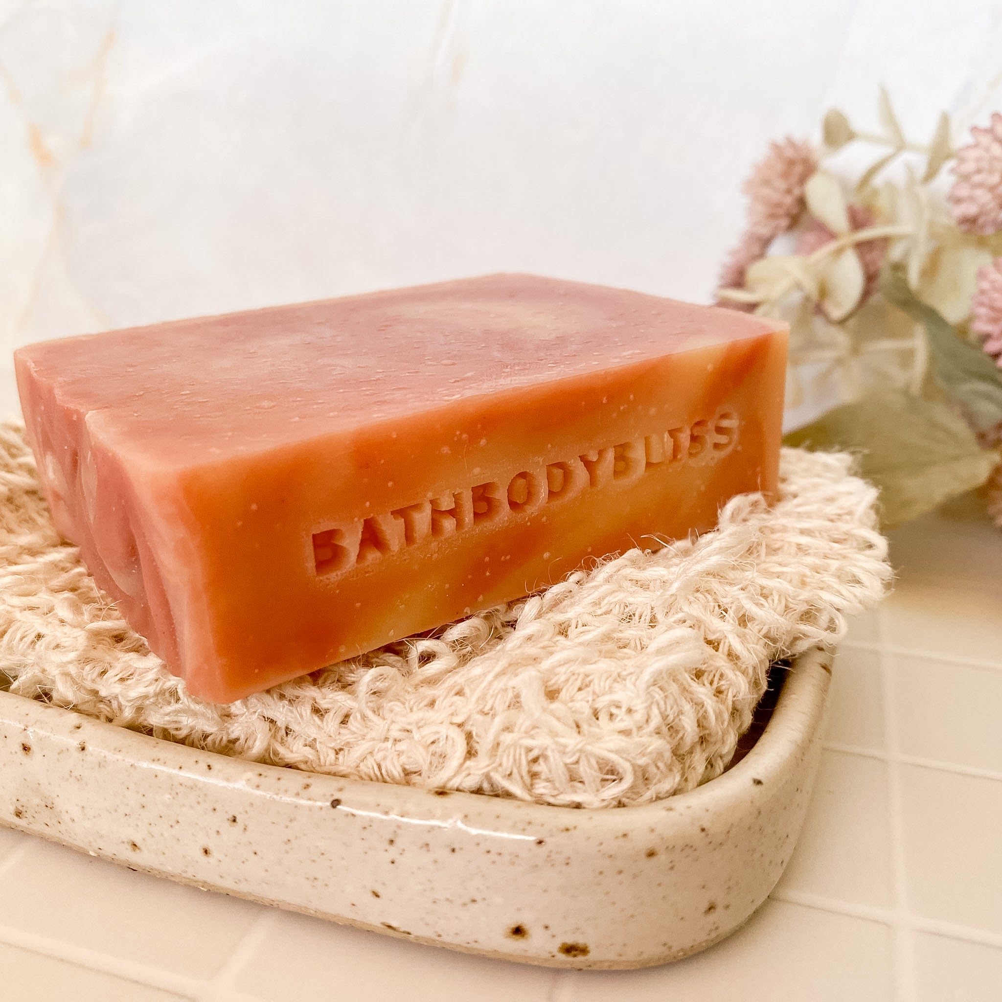 Bar of pink swirled soap with 'BathBodyBliss' branding on a natural fibre soap saver.