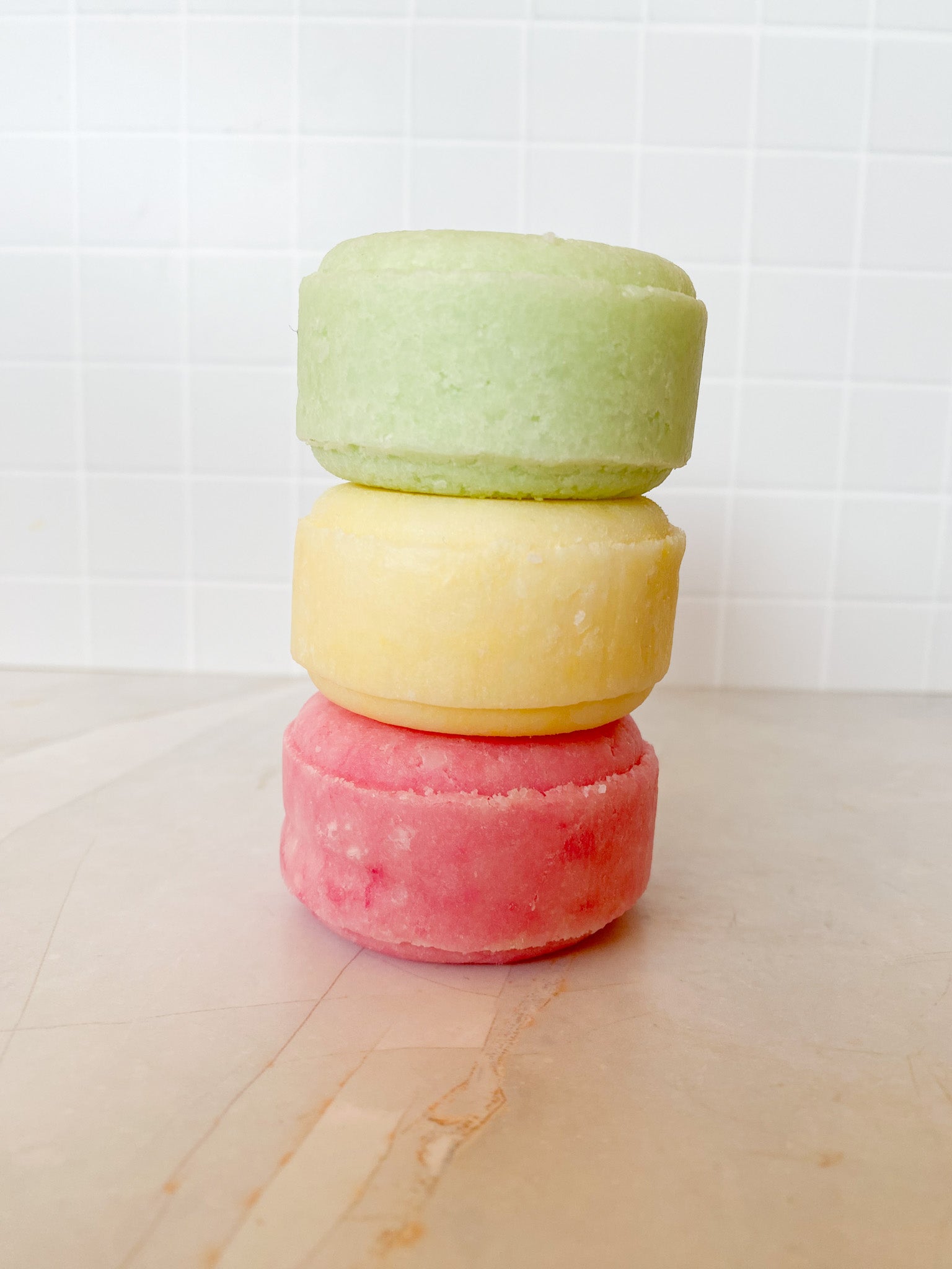 Three colorful shampoo bars stacked on a light surface with a white tiled wall background.