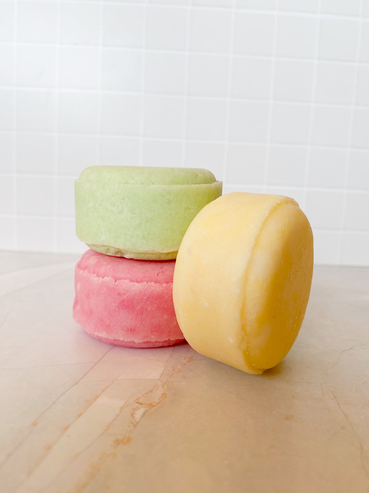 Three colorful shampoo bars stacked on a light marble surface with a white tiled wall background.