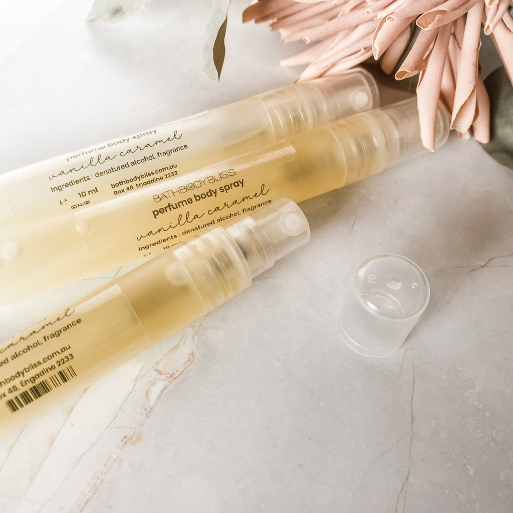 Three perfume body spray bottles with a clear liquid visible inside, placed on a surface with a decorative flower beside them, indicating the product is for scent application.