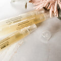 Three perfume body spray bottles with a clear liquid visible inside, placed on a surface with a decorative flower beside them, indicating the product is for scent application.