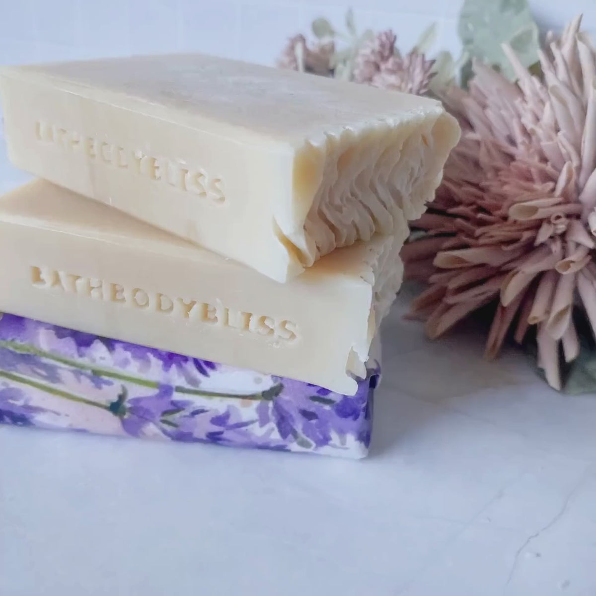 short video of three lavender and patchouli fullsize soaps with two unwrapped bars and 1 wrapped bar stacked on a pale surface with floral decor in the background