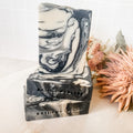 A handmade bar of vegan soap with a swirled gray and white pattern, placed on a surface with a partial view of a floral design to the side.