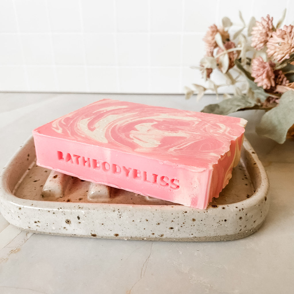 Pink and white swirled soap bar on a speckled ceramic dish with 'BathBodyBliss' branding, set against a neutral background.