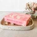 Pink and white swirled soap bar on a speckled ceramic dish with 'BathBodyBliss' branding, set against a neutral background.