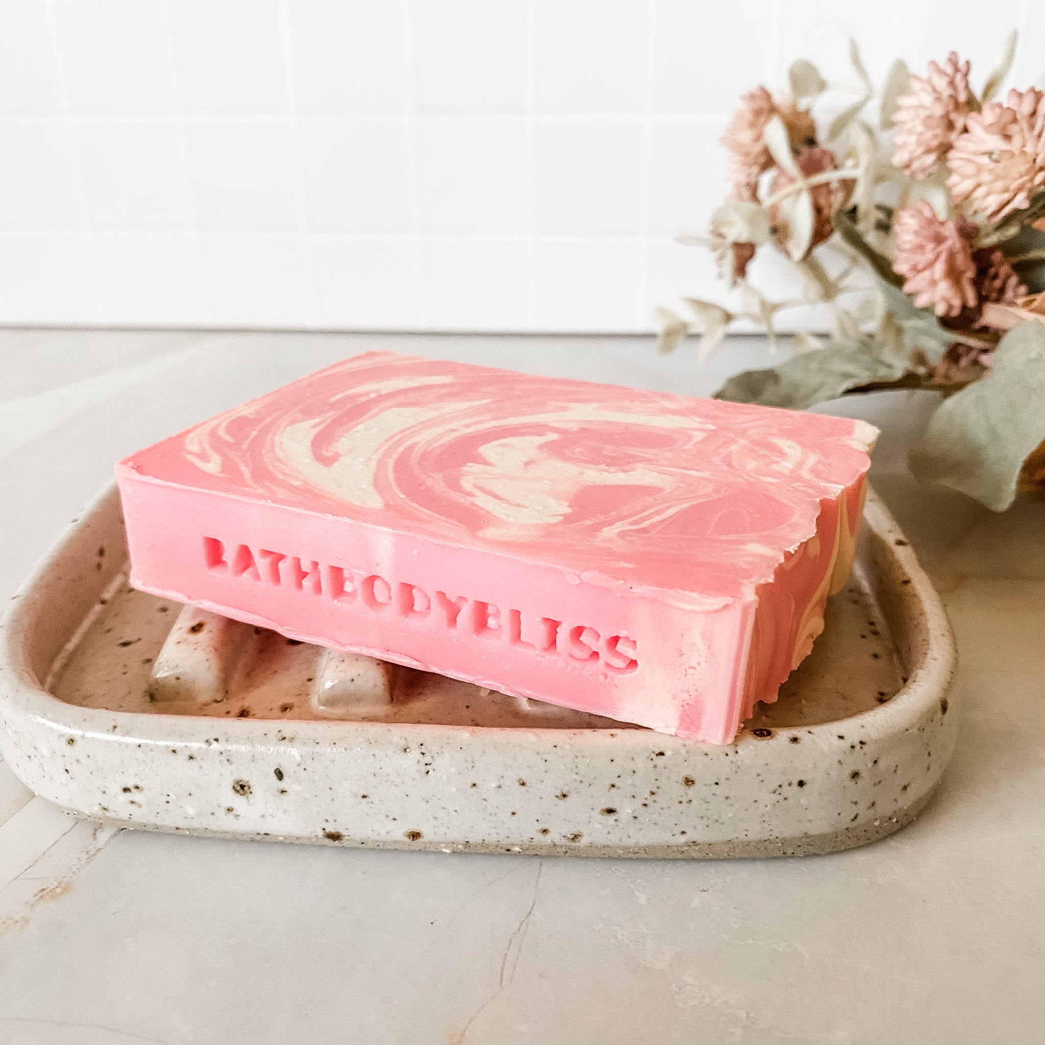 Pink and white swirled soap bar on a speckled ceramic dish with 'BathBodyBliss' branding, set against a neutral background.