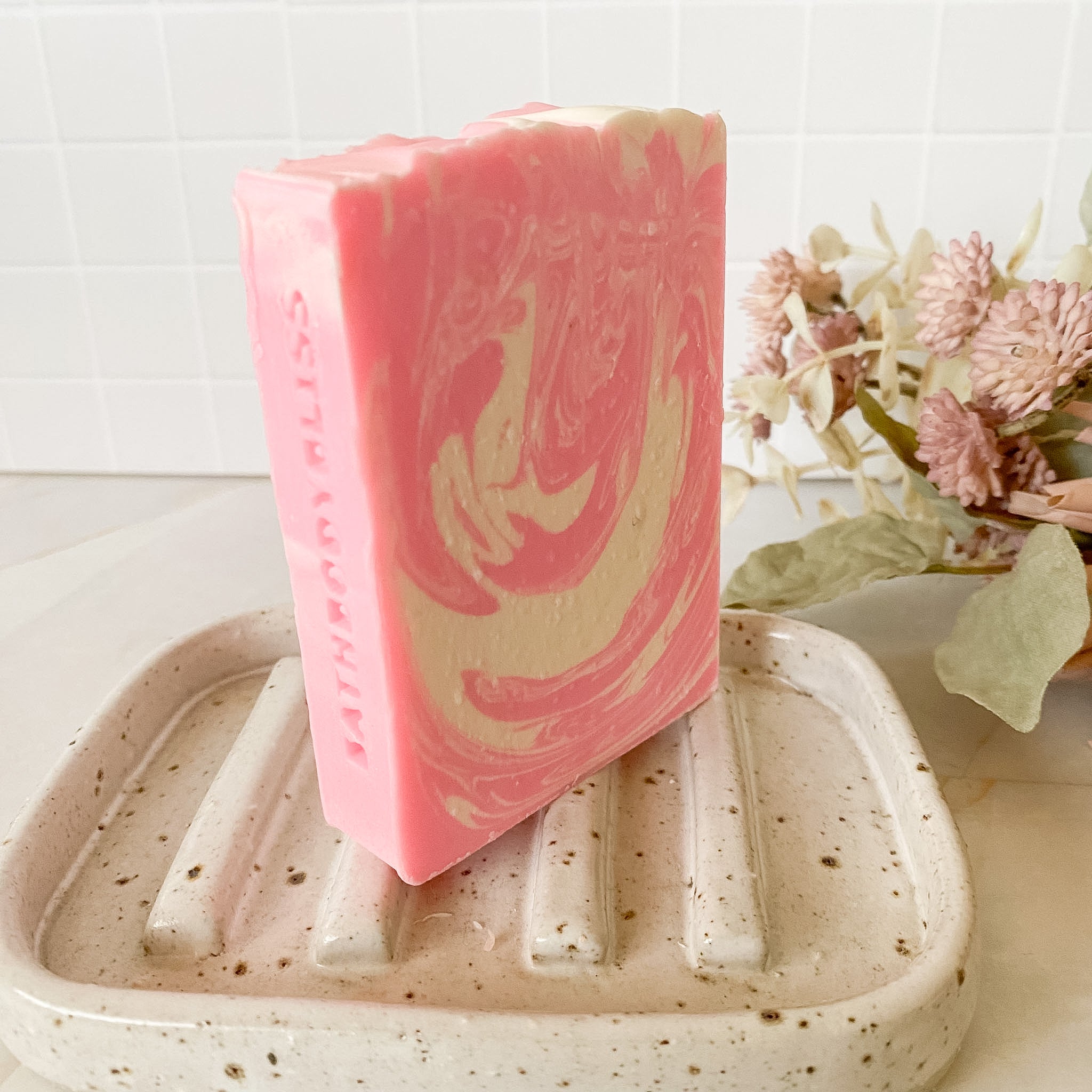 Pink and white marbled soap bar on a textured soap dish with flowers in the background