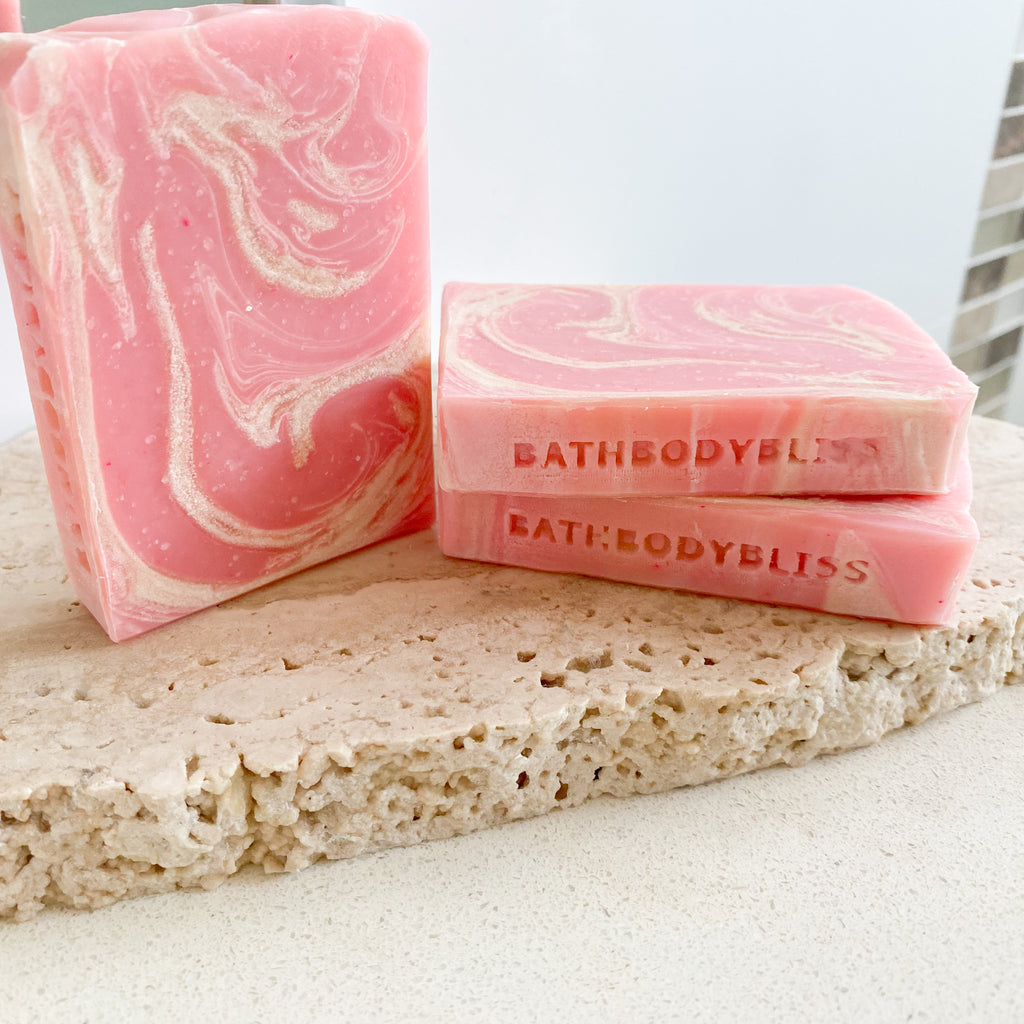 Two bars of handmade soap with a creamy pink color, one standing upright and the other lying flat, on a textured stone surface.