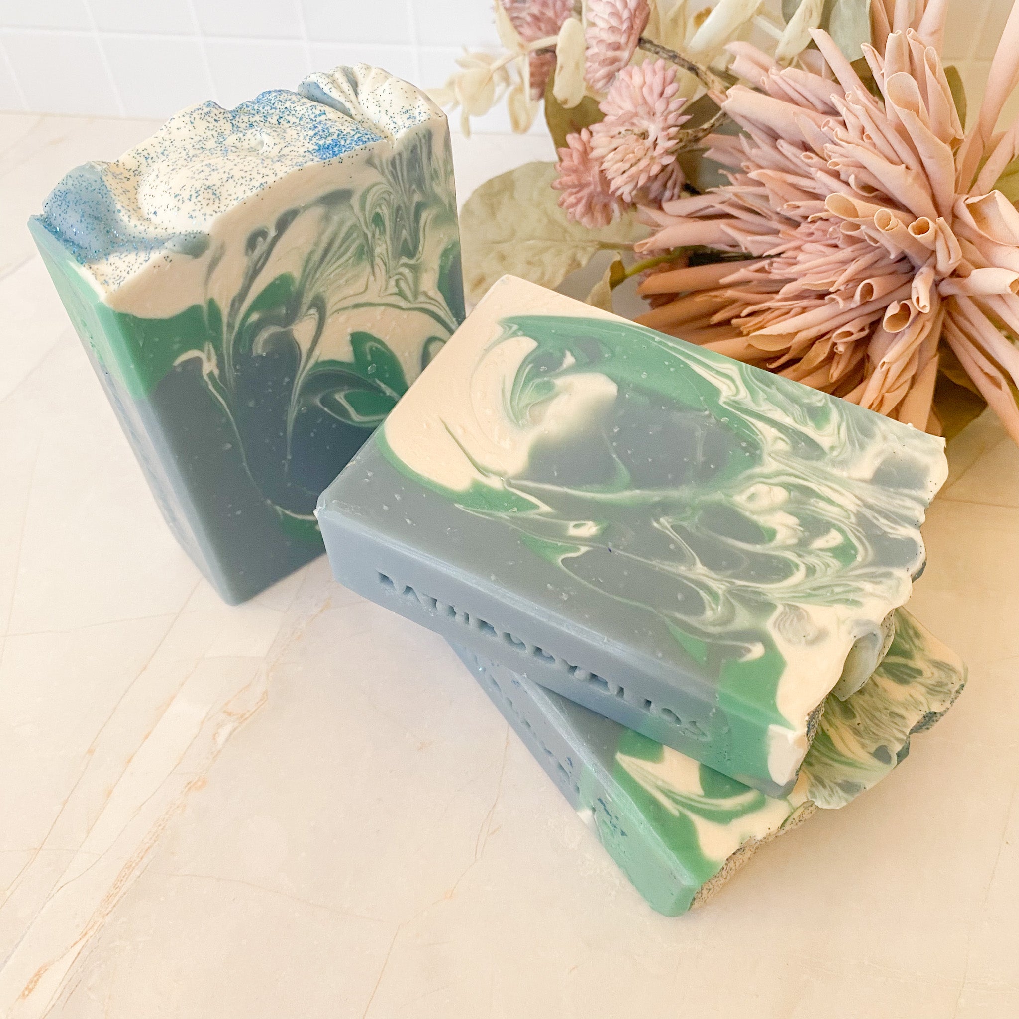 three bars of soap with blue, white and green swirls on a light surface with dried flowers in the background.