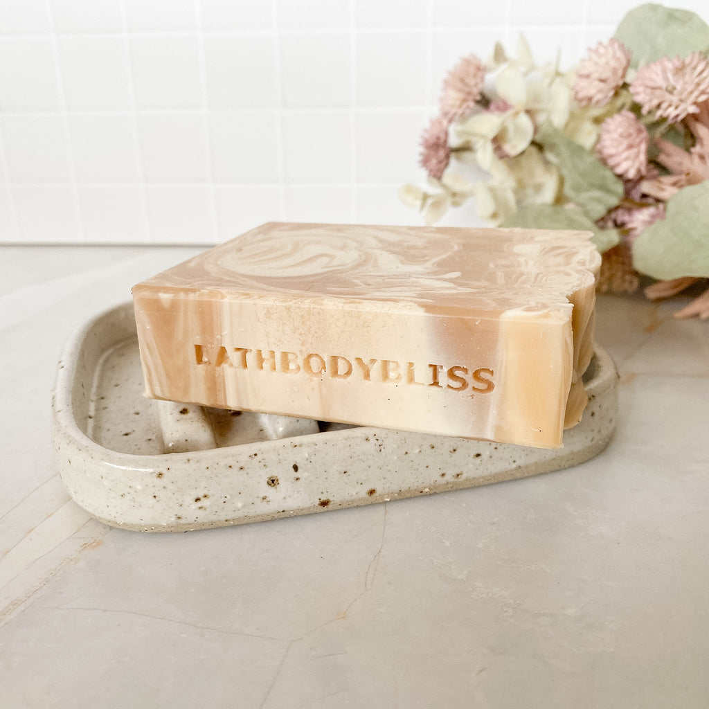 Bar of soap with 'BATHBODYBLISS' branding on a speckled stone soap dish, with flowers in the background.