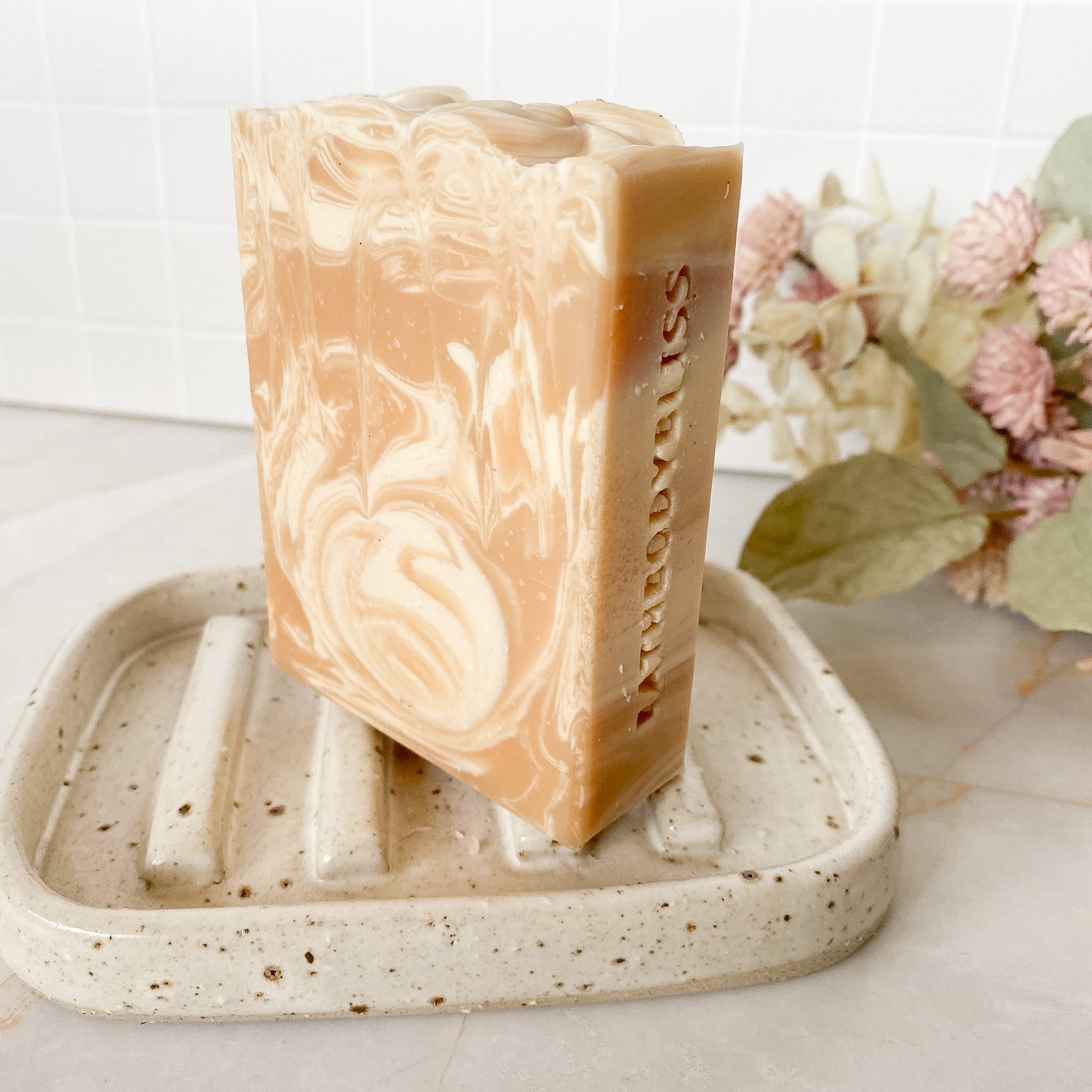 Bar of soap with 'Bath Body Bliss' branding on a speckled soap dish with flowers in the background.