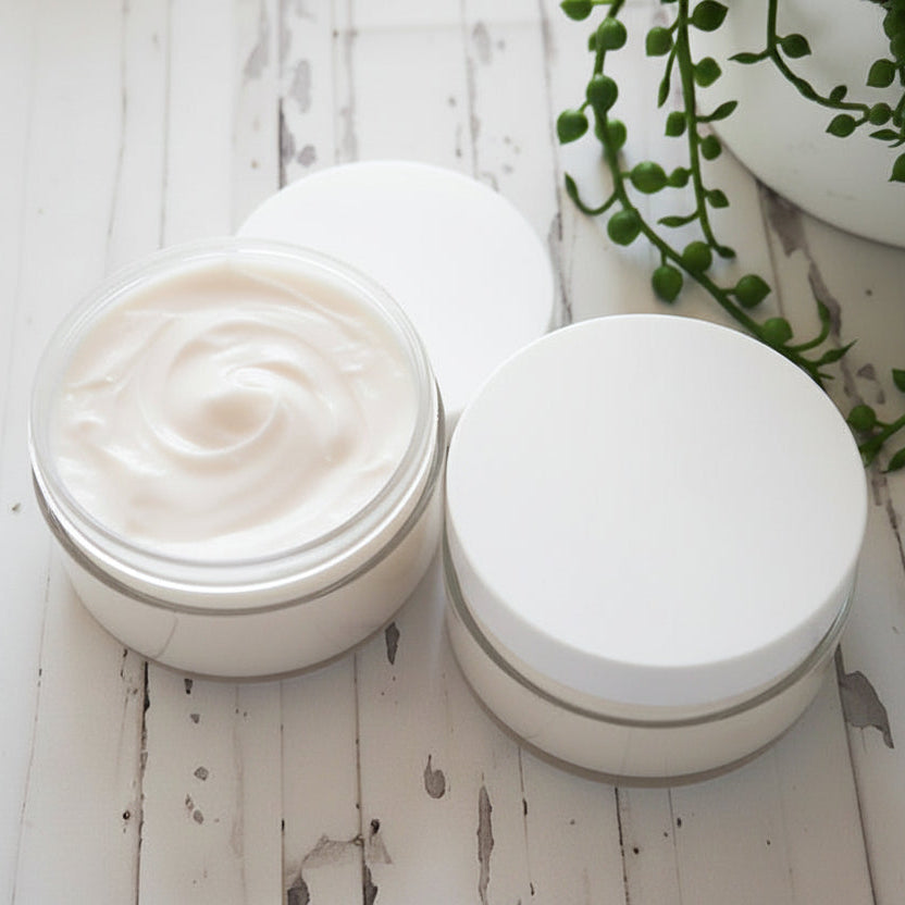 Two jars of white cream and white lids on a light wooden surface with a plant in the background